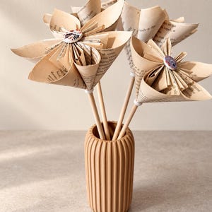 May include: Three decorative flowers crafted from vintage book pages with printed text, each featuring a central patterned button. The flowers are attached to thin wooden stems and are displayed in a vertically ribbed, cylindrical tan vase.