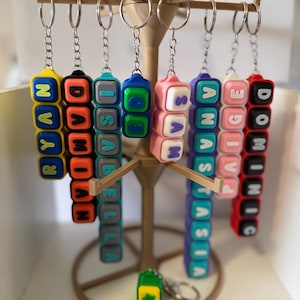 May include: A collection of colourful, rectangular silicone keychains hanging from a stand. Each keychain features raised, block-style letters spelling out names including Ryan, Damian, Isabella, Sam, Anastasia, Paige, and Dominic in various colours.