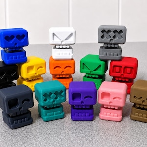 May include: A collection of small, cube-shaped skull figurines in various solid colours including blue, yellow, white, orange, green, red, black, dark grey, teal, purple, pink, and brown, each featuring distinct facial expressions and cut-out eyes.