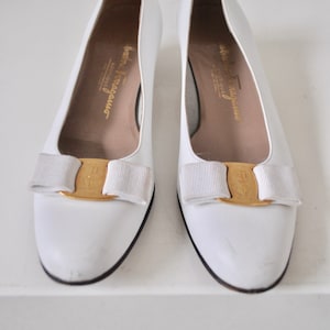 May include: A pair of white leather shoes with a gold buckle and a white bow. The shoes are made by Salvatore Ferragamo.