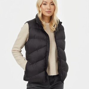 May include: A black padded gilet with a high collar and a full-length zip. The gilet is worn over a light beige jumper and paired with dark grey jeans. The model is standing in front of a white background.
