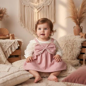 Könnte beinhalten: Ein Baby trägt ein rosa Kleid mit einer großen Schleife auf der Vorderseite, kombiniert mit einem weißen Langarmshirt. Das Baby sitzt auf einem cremefarbenen Kissen, vor einem weichen, neutralen Hintergrund.