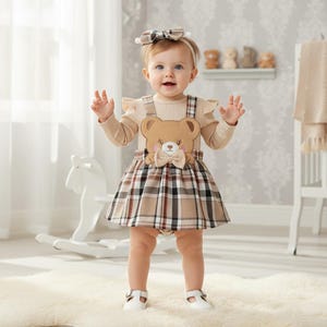 May include: A baby wearing a beige outfit with a plaid skirt and a teddy bear applique. The outfit includes a matching headband and white shoes. The baby is standing on a white rug.