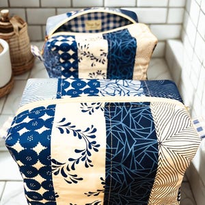 May include: Two rectangular zippered pouches featuring a patchwork design of blue and cream fabrics. The patterns include floral vines, geometric lines, and circular motifs. Each pouch has a light-colored zipper and a small blue and white checkered tab.