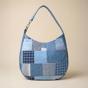 May include: A hobo-style shoulder bag crafted from a patchwork of blue and white fabric squares. The design features various patterns including gingham, stripes, herringbone, and polka dots. A small white label reads "Loryn Bags" in the center.