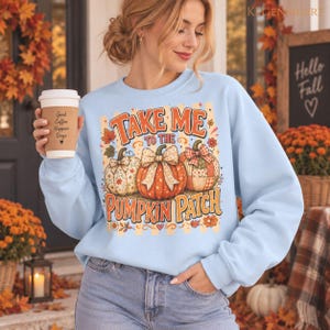 Könnte beinhalten: Ein hellblaues Langarm-Sweatshirt mit einem Grafikdruck von drei Kürbissen mit Schleifen und dem Text "TAKE ME TO THE PUMPKIN PATCH" in orangefarbenen und roten Buchstaben, verziert mit kleinen Herbstblättern und Blumen in einem Design.