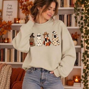 Könnte beinhalten: Ein beigefarbenes Sweatshirt mit Rundhalsausschnitt und einem Grafikdruck von vier Geistern. Der erste Geist trägt einen Hexenhut und Leopardenmuster, der zweite eine sternförmige Brille, der dritte einen Cowboyhut und Karomuster, der vierte eine schwarze Katze.