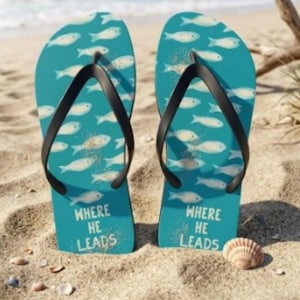 May include: A pair of turquoise flip-flops with a black thong strap. The flip-flops are decorated with white fish and the text "WHERE HE LEADS". The flip-flops are on a sandy beach with the ocean in the background.