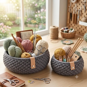 May include: Two round, dark gray crocheted baskets with light tan leather tabs and metal rivets. The baskets contain various balls of yarn in green, pink, yellow, and cream, along with wooden crochet hooks, metal scissors, and a small notebook.