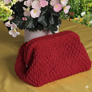 May include: A handmade, red crochet clutch bag with a textured surface. The bag is rectangular with rounded edges and a folded top. The background includes a white vase with pink flowers and green leaves.