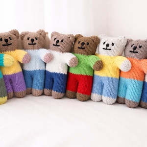Pode incluir: Uma coleção de ursos de pelúcia de malha feitos à mão em várias cores. Cada urso tem uma roupa única, incluindo suéteres e calças em tons de amarelo, azul, verde, laranja e marrom. Alguns ursos têm um detalhe de coração. Os ursos estão alinhados.