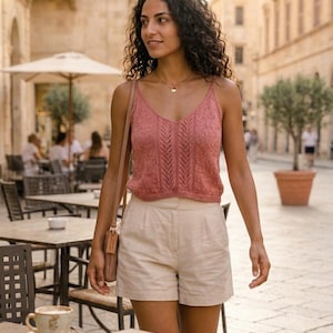 May include: A woman wearing a pink knitted camisole top and cream-colored shorts. She is walking on a cobblestone street with buildings in the background. A gift box and a cup of coffee are on a table in front of her. The text "L'ATELIER MAILLE - THE VALLETTA CAMISOLE" is at the bottom.