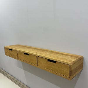 May include: A floating wooden shelf with three drawers. The shelf is made of light-colored wood and has a rectangular shape with rounded edges. Each drawer has a rectangular cutout for a handle.