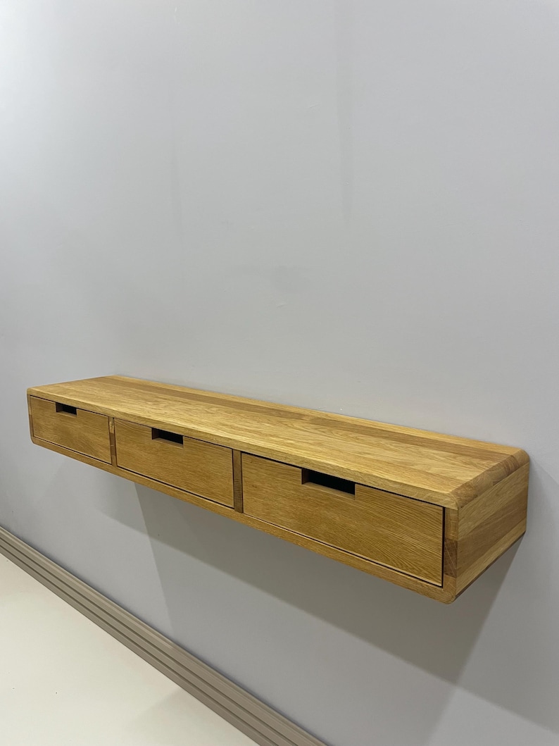 May include: A floating wooden shelf with three drawers. The shelf is made of light-colored wood and has a rectangular shape with rounded edges. Each drawer has a rectangular cutout for a handle.