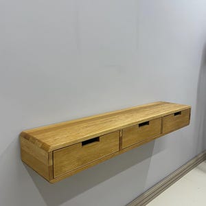 May include: A floating wooden shelf with three drawers. The shelf is made of light-colored wood and is mounted on a gray wall. Each drawer has a rectangular cutout for a handle.