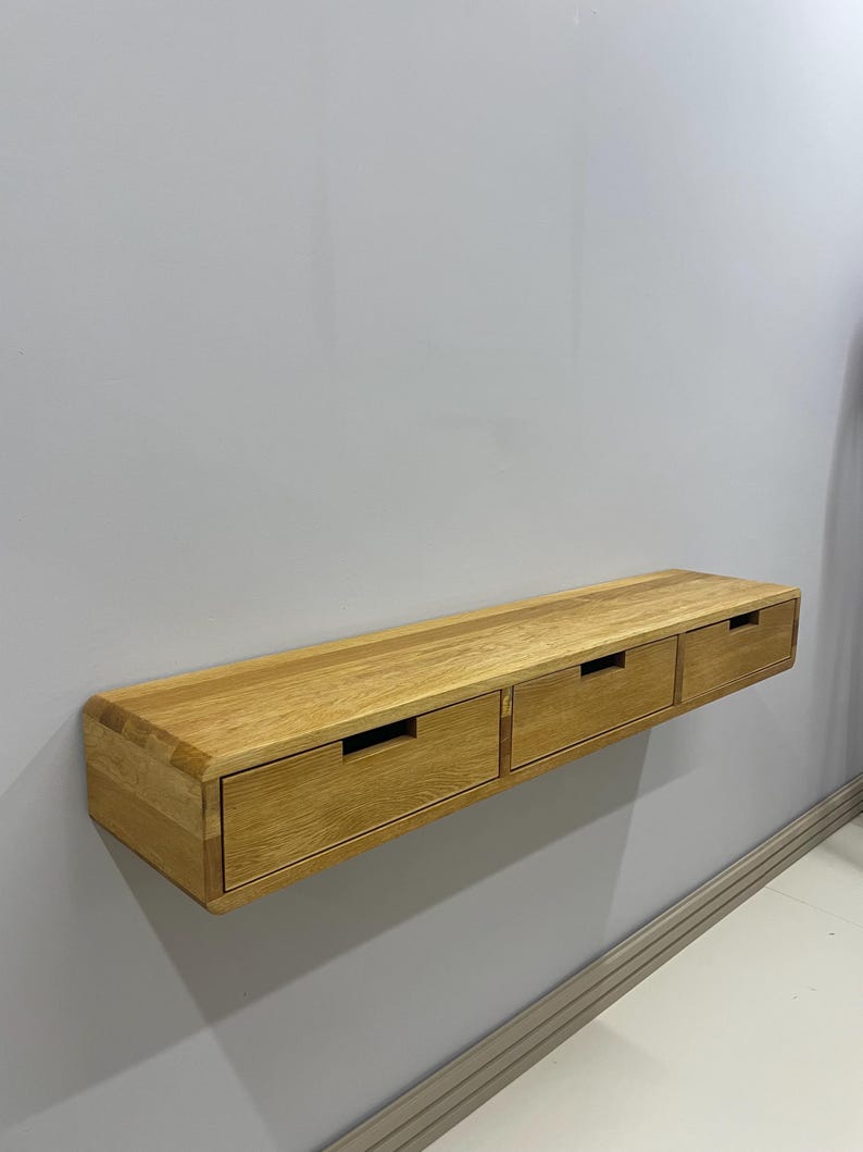 May include: A floating wooden shelf with three drawers. The shelf is made of light-colored wood and is mounted on a gray wall. Each drawer has a rectangular cutout for a handle.