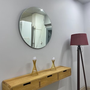 May include: A round mirror hangs above a light wood floating console with three drawers. Two gold-colored decorative objects with clear spheres sit on the console. A burgundy lampshade sits atop a dark brown tripod floor lamp.