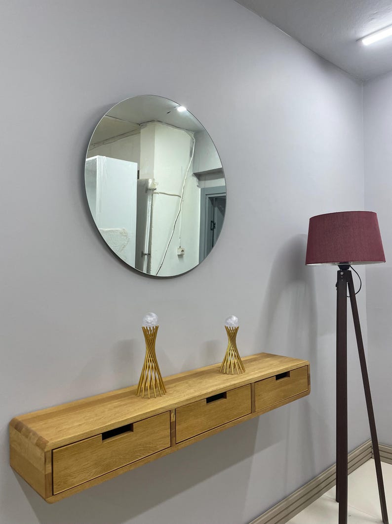 May include: A round mirror hangs above a light wood floating console with three drawers. Two gold-colored decorative objects with clear spheres sit on the console. A burgundy lampshade sits atop a dark brown tripod floor lamp.