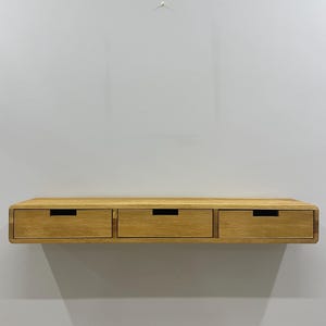 May include: A floating wooden shelf with three drawers. The shelf is made of light-colored wood and has a smooth, rounded edge. Each drawer has a dark, rectangular handle. The shelf is mounted on a light gray wall.