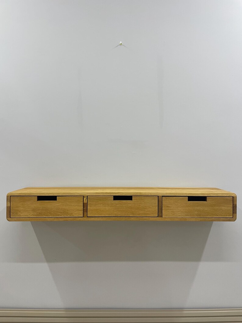 May include: A floating wooden shelf with three drawers. The shelf is made of light-colored wood and has a smooth, rounded edge. Each drawer has a dark, rectangular handle. The shelf is mounted on a light gray wall.