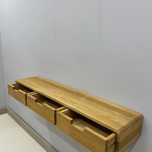 May include: A floating wooden shelf with three drawers, each partially open. The shelf is made of light-colored wood and is mounted on a gray wall. The drawers are rectangular and have a simple, modern design.