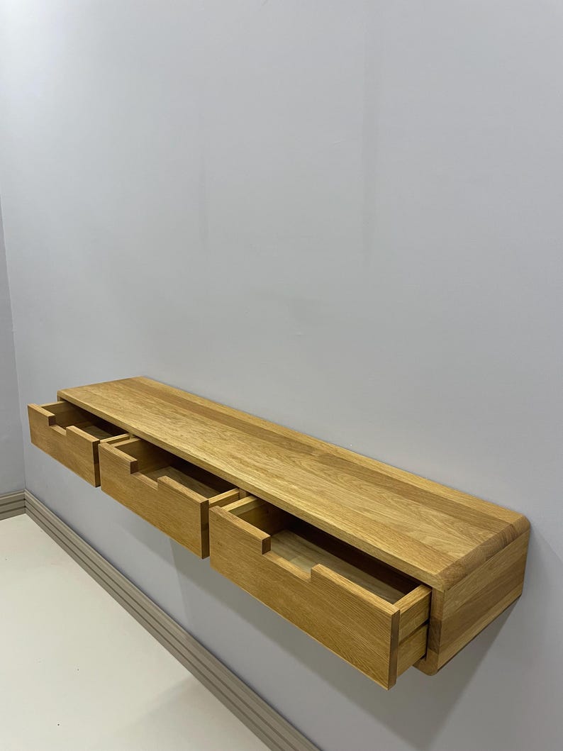 May include: A floating wooden shelf with three drawers, each partially open. The shelf is made of light-colored wood and is mounted on a gray wall. The drawers are rectangular and have a simple, modern design.