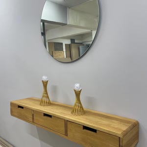 May include: A floating wooden console with three drawers, mounted on a gray wall. Above the console is a large, oval mirror. Two gold-colored decorative objects sit atop the console. The wood has a natural finish.