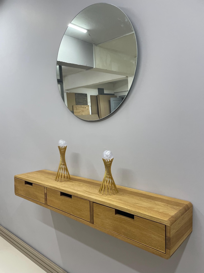 May include: A floating wooden console with three drawers, mounted on a gray wall. Above the console is a large, oval mirror. Two gold-colored decorative objects sit atop the console. The wood has a natural finish.