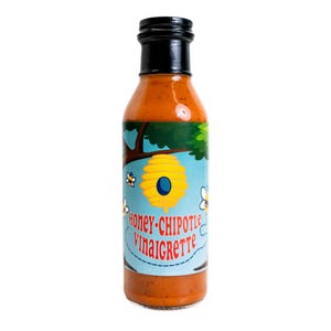 May include: A clear glass bottle filled with orange liquid, topped with a black cap. The label features a light blue background with a yellow beehive, small bees, and the red text "HONEY-CHIPOTLE VINAIGRETTE" printed in a stylized font.