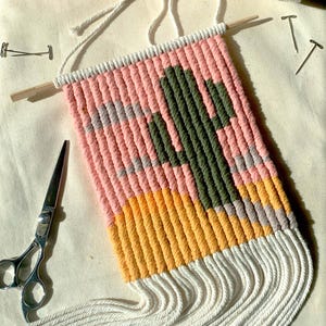 May include: A handmade macrame wall hanging featuring a cactus design. The piece uses pink, yellow, gray, and green cords, with a wooden dowel at the top and white fringe at the bottom. Scissors, pins, and yarn are also visible.