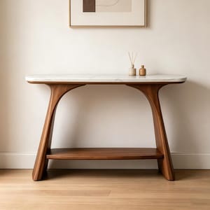Puede incluir: Una consola moderna de mediados de siglo con una tapa de mármol blanco y una base de madera oscura. La mesa tiene un diseño curvo con un estante inferior para almacenamiento. Dos pequeñas botellas decorativas están sobre la mesa. La mesa mide unos 152 cm de largo.