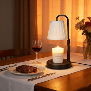 Könnte beinhalten: Eine brennende Kerze mit dem Wort "FRAGRANT" auf dem Etikett steht unter einem weißen Lampenschirm. Ein Glas Rotwein und ein Teller mit Steak stehen auf dem Tisch. Die Lampe ist schwarz und hat einen gebogenen Arm.