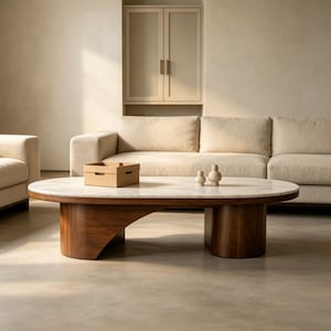 Puede incluir: Una mesa de centro ovalada con tapa de mármol y base de madera oscura. La base tiene un diseño curvo en un lado y un soporte cilíndrico en el otro. Dos pequeños objetos decorativos de color claro y una pequeña caja de cartón están sobre la mesa.