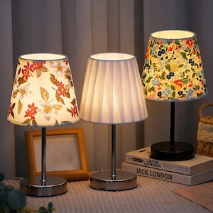 May include: Three decorative table lamps with different patterned shades. One shade features a floral design with red and white flowers, another is pleated white, and the third has a colorful floral pattern. Each lamp has a metal base.