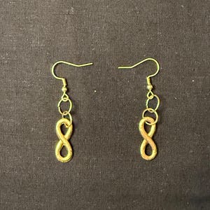 Puede incluir: Un par de pendientes dorados. Cada pendiente presenta un gancho, una pequeña cadena y un dije con forma de ocho. Los pendientes se muestran sobre un fondo oscuro.