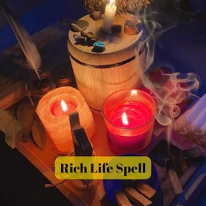 May include: A candlelit scene featuring a wooden barrel, a pink salt candle, and a red candle in a glass. The text "Rich Life Spell" is displayed on a yellow banner. Other items include a feather, incense, and small stones.