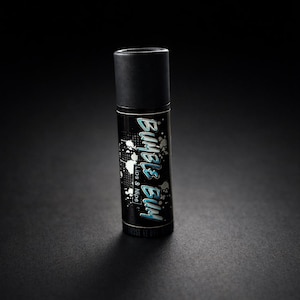 Puede incluir: Un producto cilíndrico negro con una tapa negra. La etiqueta del producto presenta el texto "Bubble Bum" en una fuente estilizada de color azul claro, con las palabras "Lips & Nips" debajo. También hay diseños blancos en forma de salpicaduras.