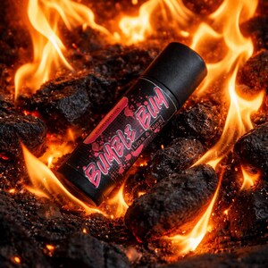 Puede incluir: Un tubo de bálsamo labial negro con el texto "Bumble Bum Lips & Nips" en rojo y blanco, sobre un fondo de brasas y llamas. La palabra "canela" también es visible en el tubo.