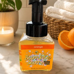Könnte beinhalten: Eine quadratische, durchsichtige Kunststoffflasche mit orangefarbener Flüssigseife und einem schwarzen Pumpspender. Das Etikett zeigt den Text "orange", "Bumble Bum", "tallow soap" und "Net Wt. 9oz (266.16mL)" sowie eine Biene und bunte Blasen.