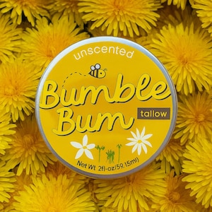 Op de afbeelding: Een ronde, gele blik van "Bumble Bum" talk, met het label "ongeparfumeerd", met een illustratie van een bij. De blik is omgeven door een achtergrond van heldergele paardenbloem bloemen. Het nettogewicht is 59,15 ml.
