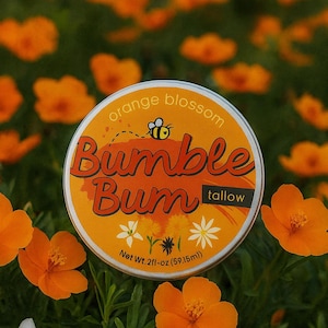 Può includere: Un barattolo rotondo, arancione e giallo di sego "Bumble Bum", con testo "orange blossom" e un'illustrazione di un'ape. Il barattolo è circondato da fiori arancioni e fogliame verde. Il peso netto è di 59,15 ml.