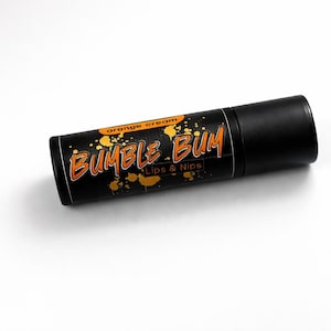 Puede incluir: Tubo cilíndrico negro de bálsamo labial "Bumble Bum". El tubo tiene texto naranja que dice "orange cream" y "Lips & Nips". El producto está sobre un fondo blanco.