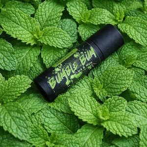 Puede incluir: Un tubo negro de bálsamo labial "Bumble Bum Lips & Nips" con texto verde, sobre un lecho de hojas frescas de menta verde. La etiqueta también incluye la palabra "peppermint".