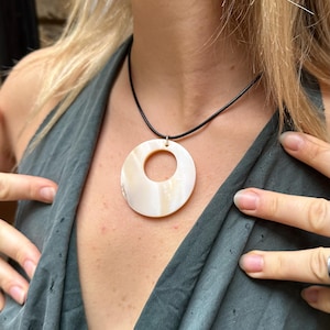 May include: A cream-coloured, circular pendant necklace with a shell-like design. The pendant hangs from a thin, black cord. The necklace is worn against a dark green top.
