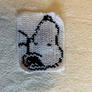 Peut inclure: Un carré blanc crocheté avec le visage d'un personnage de dessin animé noir. Cet article fait main est probablement un dessous de verre ou une petite pièce décorative, avec un design simple mais reconnaissable. Le fond est une surface texturée jaune clair.