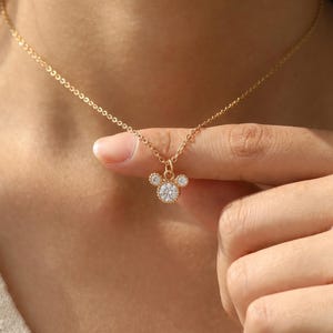 K&ouml;nnte beinhalten: Eine goldfarbene Halskette mit einer feinen Kette und einem Anh&auml;nger in Form einer Mauskopf-Silhouette. Der Anh&auml;nger ist mit drei klaren, rund geschliffenen Steinen besetzt, wobei ein gr&ouml;&szlig;erer Mittelstein und zwei kleinere Steine die Ohren bilden.
