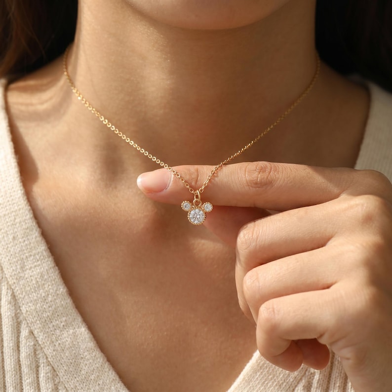 K&ouml;nnte beinhalten: Eine goldfarbene Halskette mit einer feinen Kette und einem Anh&auml;nger in Form einer Mauskopf-Silhouette. Der Anh&auml;nger ist mit drei klaren, rund geschliffenen Steinen besetzt, wobei ein gr&ouml;&szlig;erer Mittelstein und zwei kleinere Steine die Ohren bilden.