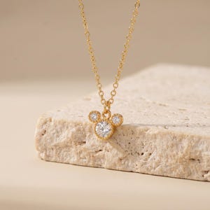 K&ouml;nnte beinhalten: Eine goldfarbene Halskette mit einem Anh&auml;nger in Form eines Mauskopfes, der mit drei runden, klaren, facettierten Steinen in Metallfassung besetzt ist. Der Anh&auml;nger h&auml;ngt an einer zarten goldfarbenen Kette mit kleinen, gleichm&auml;&szlig;igen Gliedern.