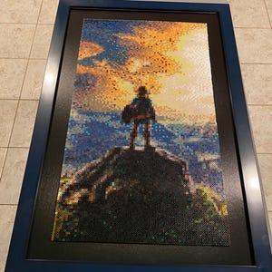 May include: Framed pixel art depicting a figure standing on a mountain, silhouetted against a sunset. The artwork is composed of small, colorful beads, creating a mosaic effect. The frame is a dark blue, adding a contrasting border to the vibrant image.