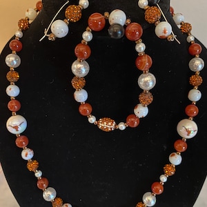 May include: A jewellery set comprising a necklace, bracelet, and earrings. The necklace and bracelet feature red, white, silver, and orange beads. The earrings have a similar design. The jewellery is displayed on a black surface.